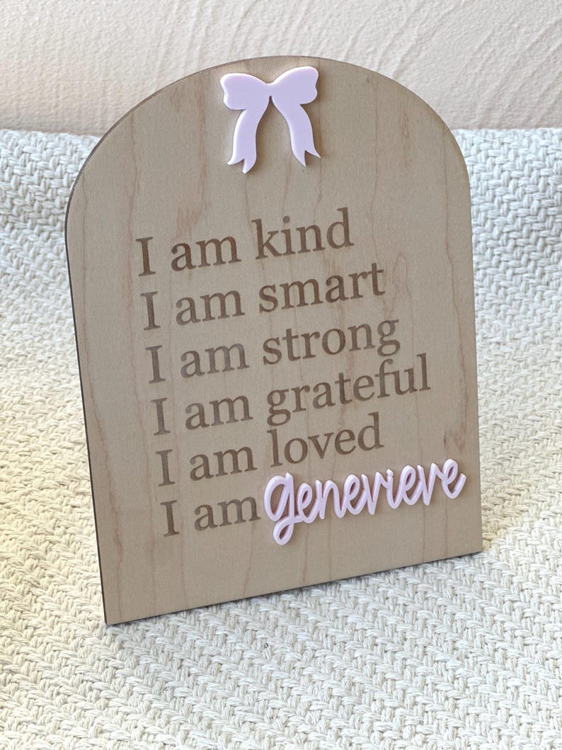 May include: A wooden sign with a pink bow at the top. The sign has the text "I am kind, I am smart, I am strong, I am grateful, I am loved, I am Genevieve" written on it.