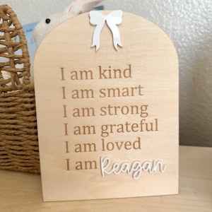 May include: A wooden sign with a white bow on top. The sign has the text "I am kind, I am smart, I am strong, I am grateful, I am loved, I am Reagan" engraved on it.