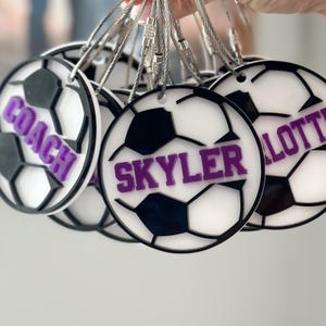 Op de afbeelding: Een set witte voetbalvormige sleutelhangers met zwarte stiksels en paarse letters. De sleutelhangers hebben de namen "Coach", "Skyler" en "Lottie".