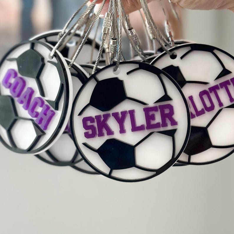 Op de afbeelding: Een set witte voetbalvormige sleutelhangers met zwarte stiksels en paarse letters. De sleutelhangers hebben de namen "Coach", "Skyler" en "Lottie".