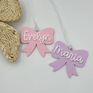 May include: Two pink and purple bow-shaped luggage tags with clear strings. The pink tag says "Evelyn" and the purple tag says "Maria".