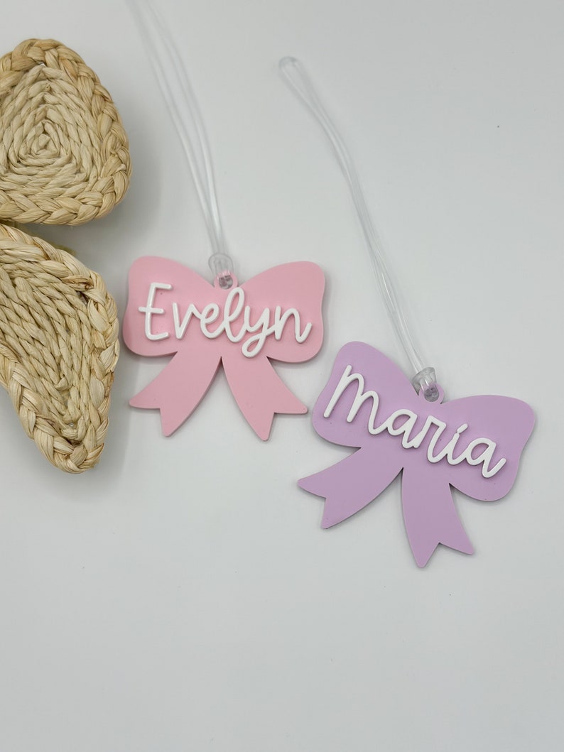 May include: Two pink and purple bow-shaped luggage tags with clear strings. The pink tag says "Evelyn" and the purple tag says "Maria".