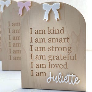 May include: Wooden plaque with a white bow on top. The plaque has the text "I am kind, I am smart, I am strong, I am grateful, I am loved, I am Juliette" engraved on it.