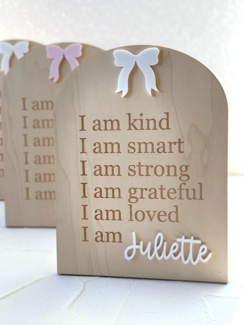 May include: Wooden plaque with a white bow on top. The plaque has the text "I am kind, I am smart, I am strong, I am grateful, I am loved, I am Juliette" engraved on it.