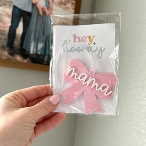May include: Pink acrylic bow with the word "mama" in white lettering. The bow is packaged in a clear plastic bag with a white card that says "hey hooray".