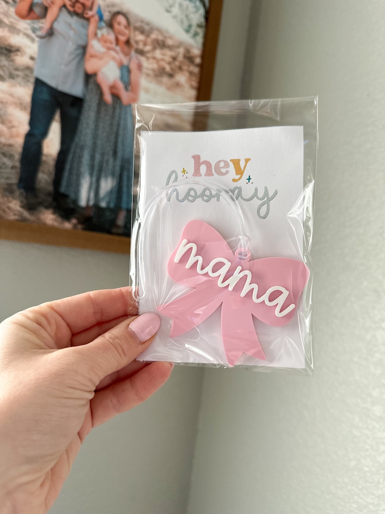 May include: Pink acrylic bow with the word "mama" in white lettering. The bow is packaged in a clear plastic bag with a white card that says "hey hooray".