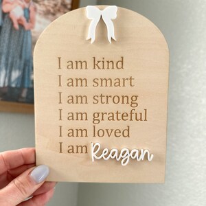 May include: A wooden sign with a white bow at the top. The sign has the text "I am kind, I am smart, I am strong, I am grateful, I am loved, I am Reagan" engraved on it.