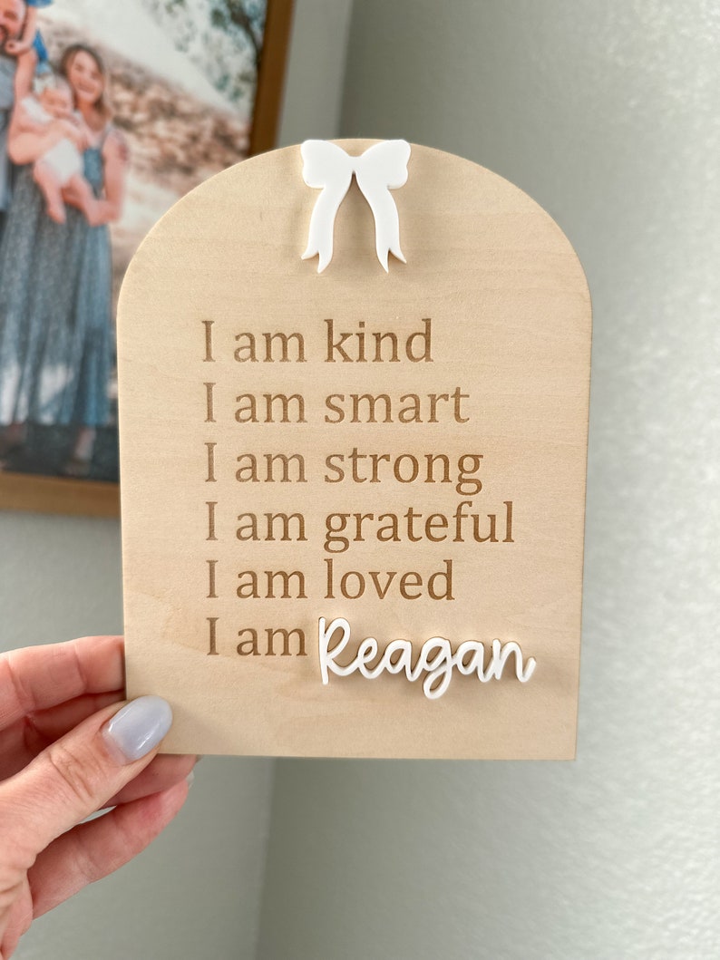 May include: A wooden sign with a white bow at the top. The sign has the text "I am kind, I am smart, I am strong, I am grateful, I am loved, I am Reagan" engraved on it.