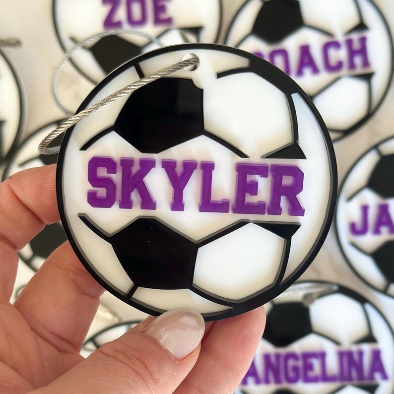 Op de afbeelding: Witte en zwarte voetbalvormige bagagelabel met de naam "Skyler" in paars gedrukt.