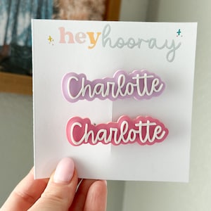 May include: Two pink and purple plastic hair clips with the name "Charlotte" written in white on each clip. The clips are on a white card with the text "hey hooray" written in colorful letters.