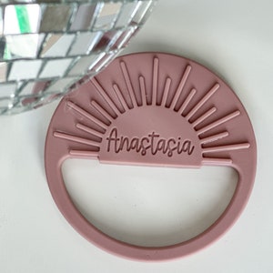 May include: A dusty rose-colored round coaster with the name "Anastasia" in a script font. The coaster features a sunburst design and a handle. A disco ball is partially visible in the background.