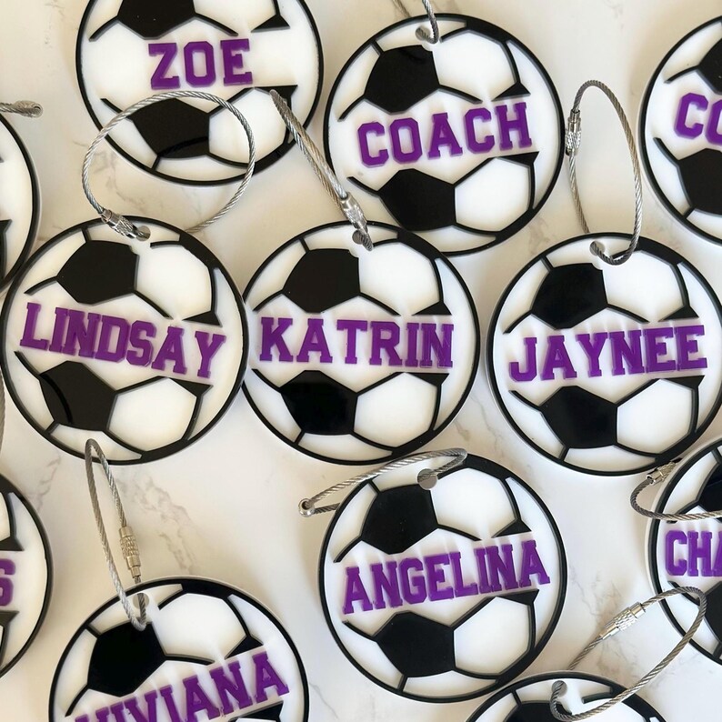 Op de afbeelding: Een verzameling witte en zwarte voetbalvormige bagagelabeltjes met paarse tekst. De labeltjes hebben namen zoals Zoe, Coach, Lindsay, Katrin, Jaynee, Angelina, Viviana en Cha.
