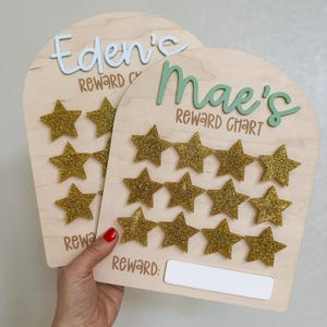 May include: Two wooden reward charts with gold glitter stars. The chart on the left says "Eden's Reward Chart" and the chart on the right says "Mae's Reward Chart".
