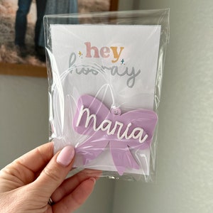 May include: A purple bow with the name "Maria" written in white lettering. The bow is packaged in a clear plastic bag with a white card that says "hey hooray".
