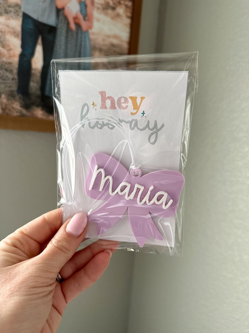May include: A purple bow with the name "Maria" written in white lettering. The bow is packaged in a clear plastic bag with a white card that says "hey hooray".