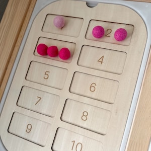 May include: A wooden counting board with pink felt balls. The board has numbers 1 through 10 printed on it. There are 3 pink felt balls in the space for number 3, 2 pink felt balls in the space for number 2, and 1 pink felt ball in the space for number 1.