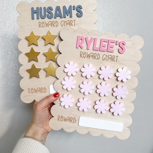 May include: Two wooden reward charts with scalloped edges. The top chart is for Husam and features gold stars. The bottom chart is for Rylee and features pink flowers. Both charts have a blank space for a reward.