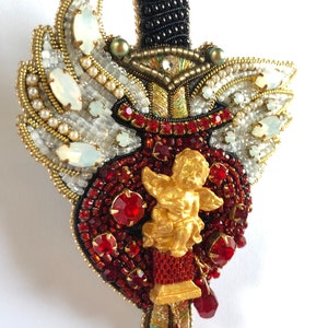 Könnte beinhalten: Eine Brosche aus Perlen in Form eines Herzens, das von einem Schwert durchbohrt wird. Das Herz ist mit roten Strasssteinen und einem goldenen Cherub verziert. Das Schwert ist mit schwarzen, weißen und goldenen Perlen verziert.