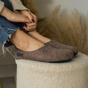 May include: A pair of brown felt slippers with black soles. The slippers are on a white fluffy surface.