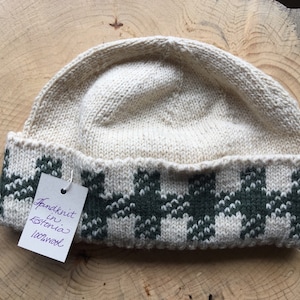 May include: A cream-coloured knitted hat with a dark green geometric pattern on the brim. The hat is hand-knitted in Estonia from 100% wool. A small tag is attached to the hat.