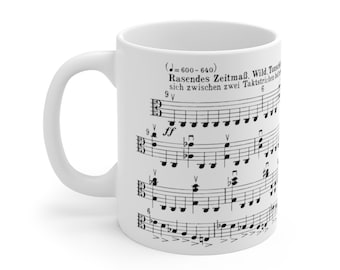 May include: White ceramic mug featuring a black and white musical score printed on it. The score includes the text "Rasendes Zeitmaß. Wild. Tonstück" and "sich zwischen zwei Taktstrichen bewegt".
