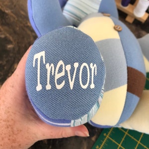 May include: A patchwork teddy bear in blue, brown, and cream. The bear's paw displays the name "Trevor" in white lettering. The bear is made from various fabrics and has button details. A handcrafted item.