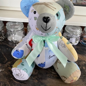 May include: A patchwork teddy bear with a black heart-shaped nose and a pale green bow. The bear's body is made of various patterned fabrics in blue, green, and beige. The bear has a grey face and ears, and the text "Buddy" is visible on one arm.