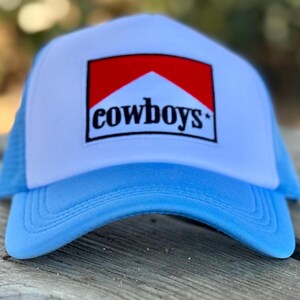 May include: A light blue trucker hat with a white front panel and a red and white logo that says "cowboys" in black letters.
