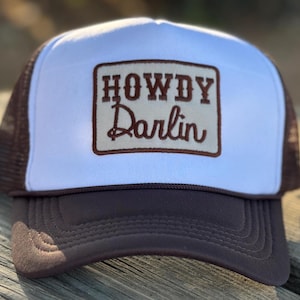 May include: A white and brown trucker hat with a brown patch that says "Howdy Darlin'" in white lettering.