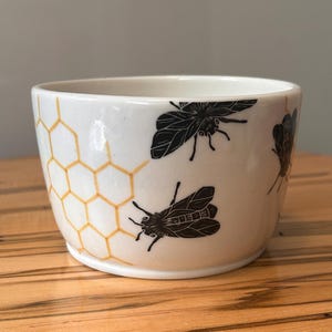 May include: A white ceramic bowl featuring a repeating pattern of black bees and a yellow honeycomb design. The bowl has a smooth, glossy finish and a rounded shape, showcasing detailed illustrations of bees in flight alongside geometric cells.