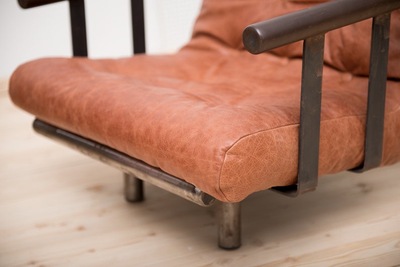 May include: A brown leather armchair with a metal frame. The chair has a simple design with a wide seat and a low back.