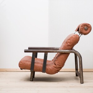May include: A brown leather armchair with a metal frame. The chair has a high back and a headrest. The chair is sitting on a wooden floor.