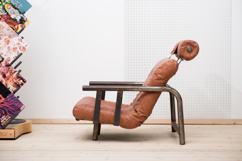 May include: A brown leather armchair with a metal frame. The chair has a high back and a headrest. The chair is sitting on a wooden floor.