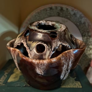 May include: A brown ceramic flower pot with white and green glaze. The pot has a round top with holes for flowers and a scalloped edge.