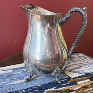 May include: A silver-coloured jug with a curved handle and three decorative feet. The jug has a wide spout and a bulbous body. The handle is ornate, and the feet have a detailed design. The jug is on a blue and white patterned surface.