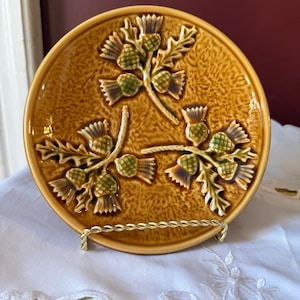 May include: A decorative, round, ceramic plate with a raised thistle design in shades of brown and green. The plate is displayed on a gold-coloured metal stand. The plate has a textured surface and a glossy finish.