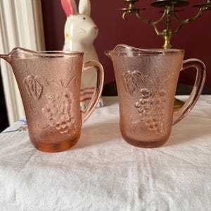 May include: Two pink glass pitchers with a textured surface and grape design. Each pitcher has a handle and a spout. The pitchers are on a white surface.