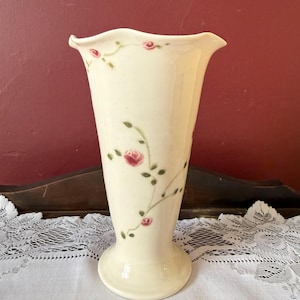 May include: Cream-colored ceramic vase with a flared top and a round base. The vase is adorned with delicate pink rose and green vine designs. The vase is approximately 8 inches tall.