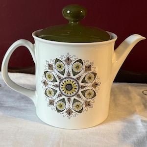 May include: White ceramic teapot with a green lid and handle. The teapot has a floral design with a sunflower in the centre, surrounded by yellow and brown accents. The spout is curved, and the handle is arched.