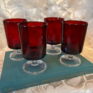 May include: Set of four ruby red glass stemmed drinking glasses with clear bases. The glasses are sitting on a teal book with gold lettering that reads "A Dog of Flanders".