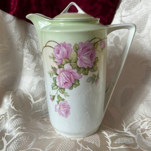 May include: A white porcelain coffee pot with a green lid and a floral design featuring pink roses. The pot has a handle and a spout.