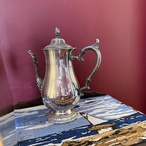 May include: A silver coffee pot with decorative features, including a detailed handle and lid. The pot is placed on a blue and white nautical-themed tablecloth. The background is a deep red wall.