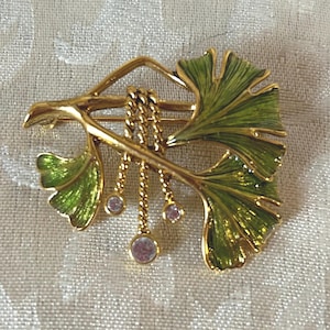 May include: Gold-coloured metal brooch in the shape of a ginkgo leaf with green enamel detailing. The brooch features three dangling chains with pink, round gemstones. The ginkgo leaf design is a classic and elegant accessory.