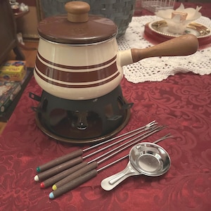 May include: A white and brown fondue pot with a wooden handle and a metal stand. The pot has two brown stripes around it. The set includes six fondue forks with wooden handles and a small metal spoon.