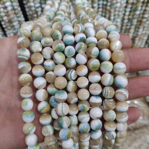 Op de afbeelding: Een streng ronde, witte, beige en blauwe iriserende schelp kralen. De kralen hebben een draaiende patroon en zijn geregen op een heldere draad.