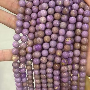 May include: Strands of round, lavender-colored beads. The beads have a smooth, polished surface with variations in color, including hints of brown and gold. These beads are strung together, suggesting they are for jewelry making or crafting.