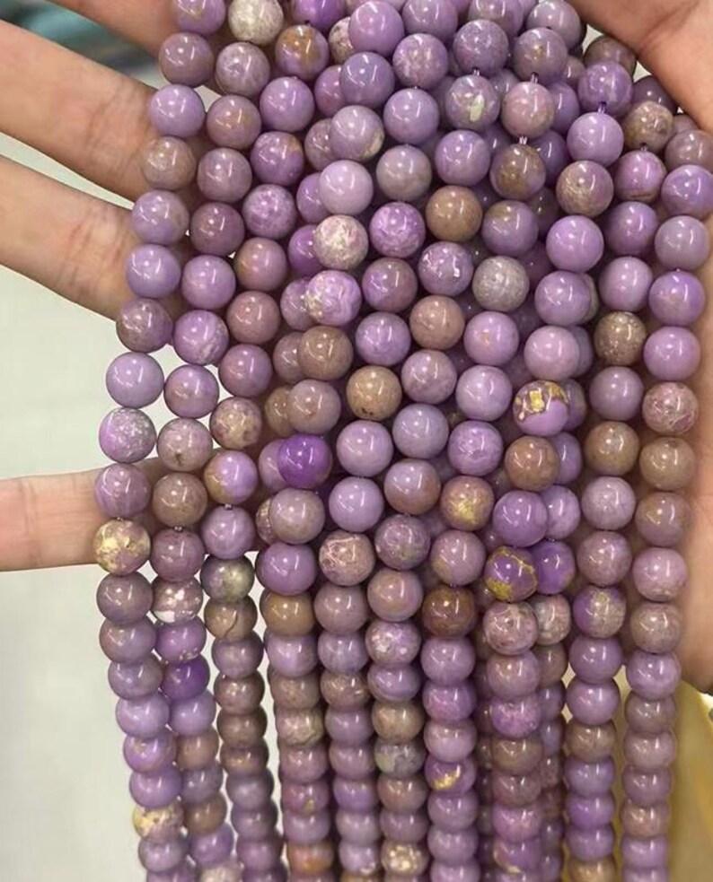 May include: Strands of round, lavender-colored beads. The beads have a smooth, polished surface with variations in color, including hints of brown and gold. These beads are strung together, suggesting they are for jewelry making or crafting.