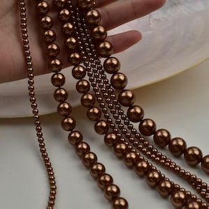 May include: Several strands of brown pearl necklaces of varying sizes are displayed. The pearls range from small seed beads to larger, round beads. The necklaces are a warm, metallic brown color, and are displayed against a white background.