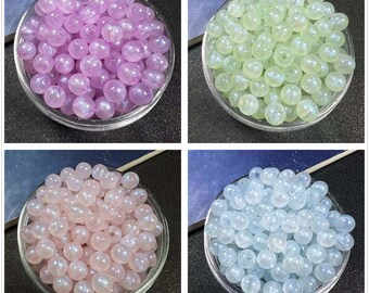 May include: Four clear glass bowls filled with iridescent beads in various pastel colors: lavender, mint green, pale pink, and light blue. The beads are round and have a shimmering, pearlescent finish, suggesting they are made of glass or a similar material.