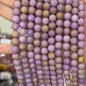 May include: Strands of round, polished beads in shades of lavender and tan. The beads are strung together, creating a textured surface. The beads are likely made of stone or glass, and are suitable for jewelry making.